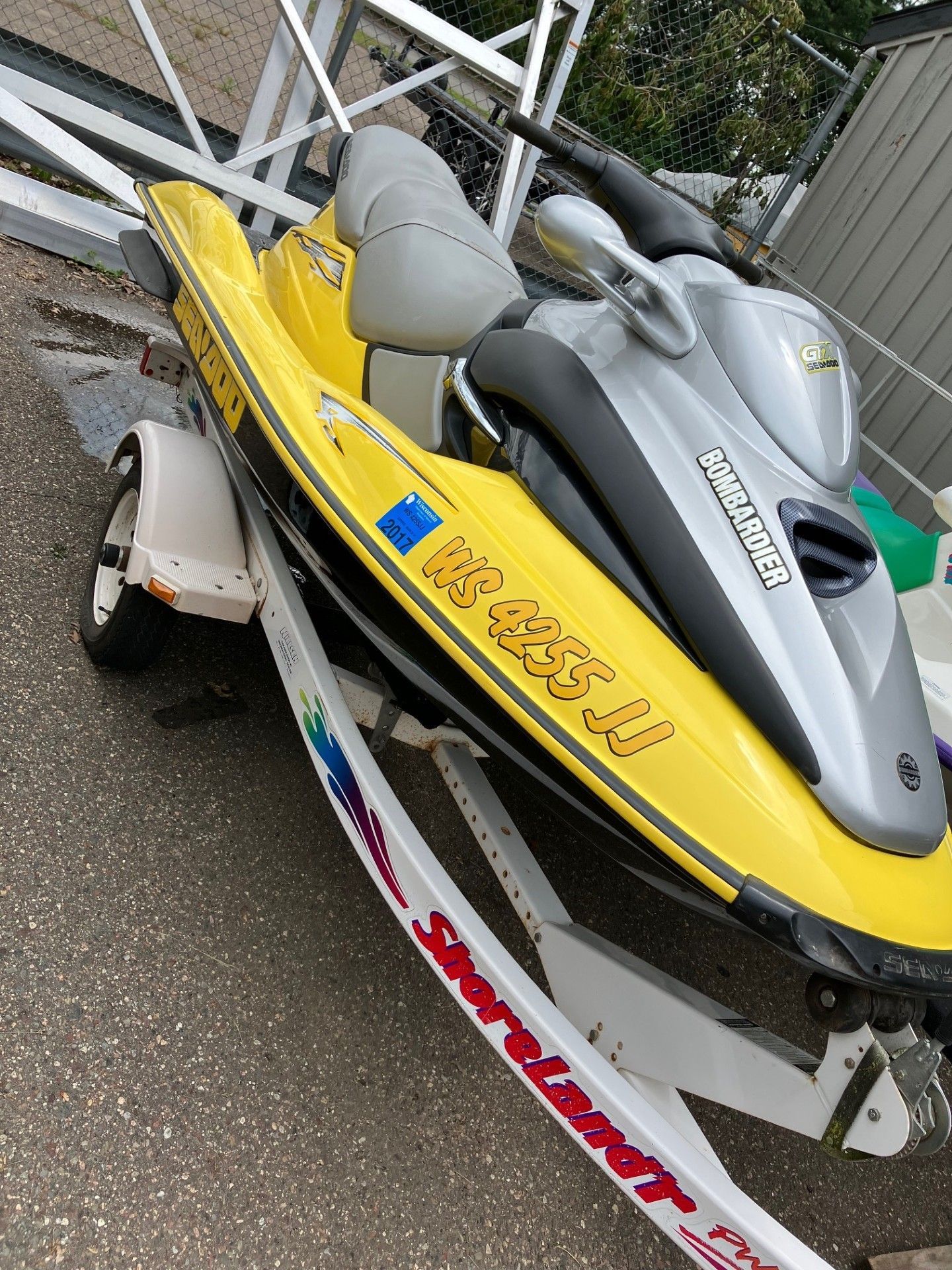 A yellow and silver Sea-Doo personal watercraft sits secured on a white ShoreLand'r boat trailer on an asphalt surface. 