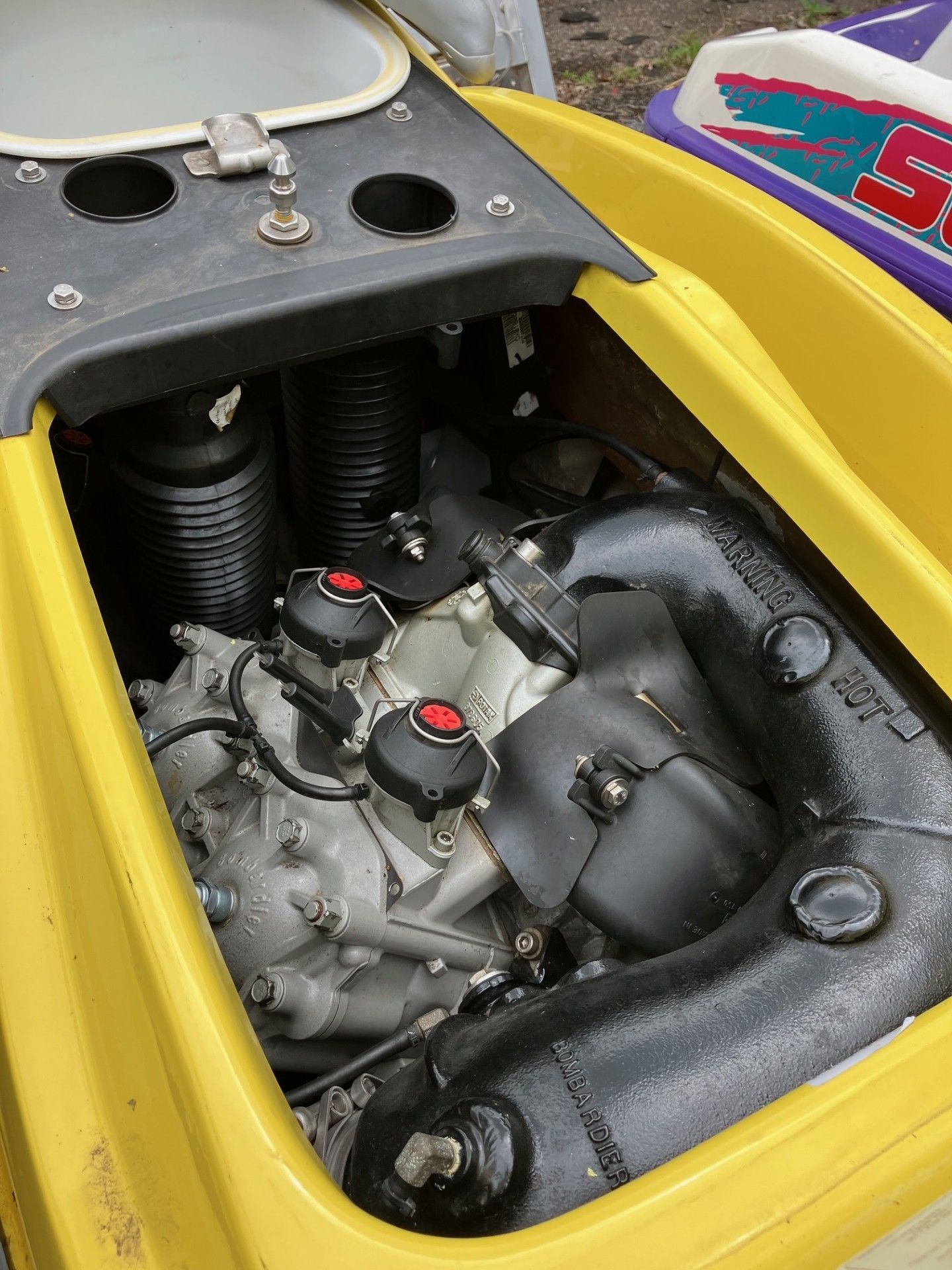An open engine compartment of a yellow personal watercraft, revealing the motor, intake tubes, and exhaust system. 