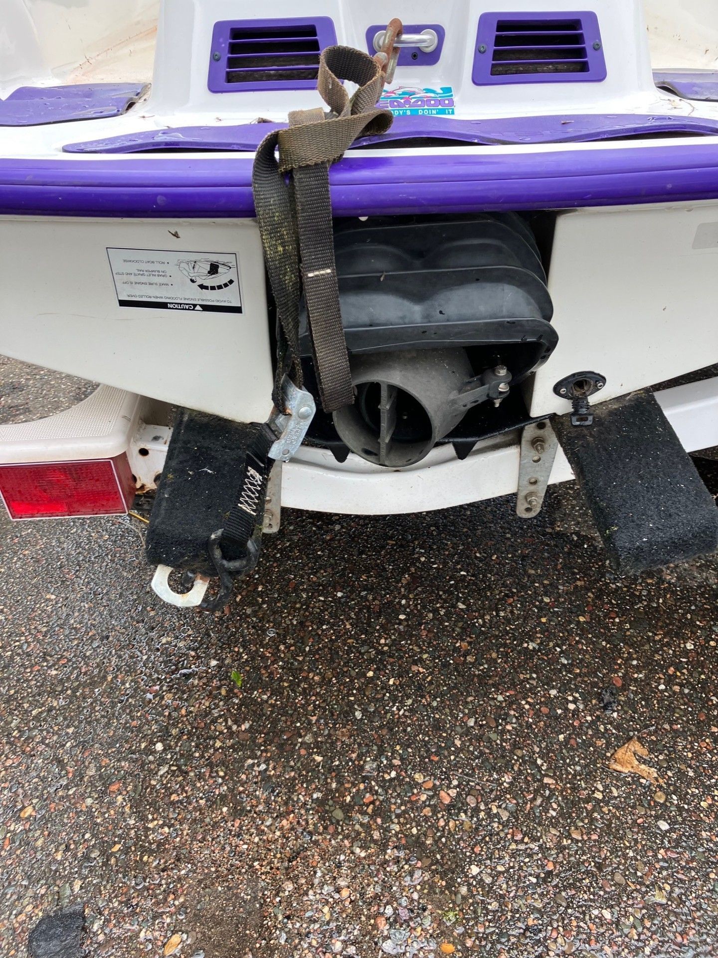 The rear view of a white and purple jet ski secured to a trailer by a brown strap, highlighting the engine exhaust nozzle. 