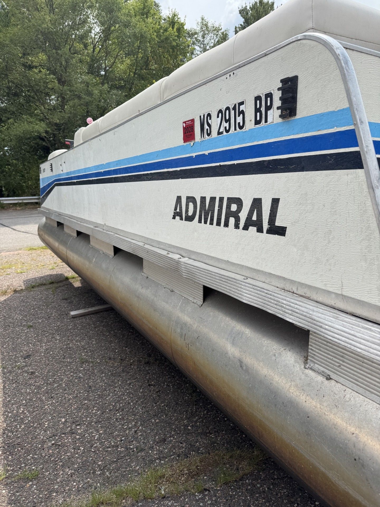 White Admiral boat with blue stripe on trailer, parked on pavement by trees