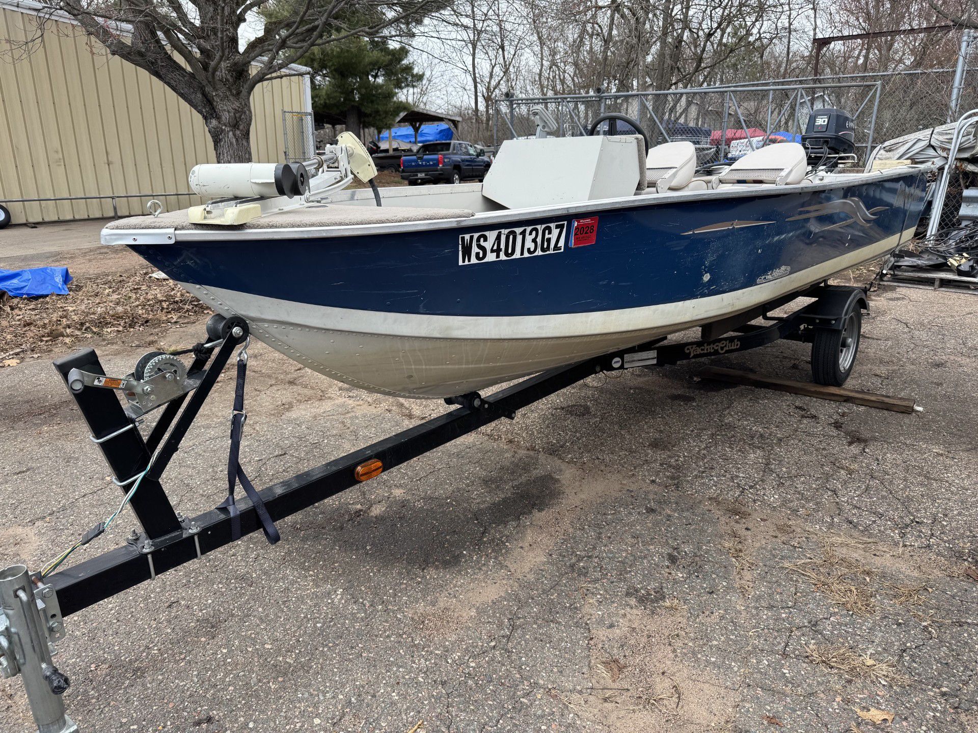 Blue fishing boat on a trailer in a gravel lot, with white seats and a small outboard motor.