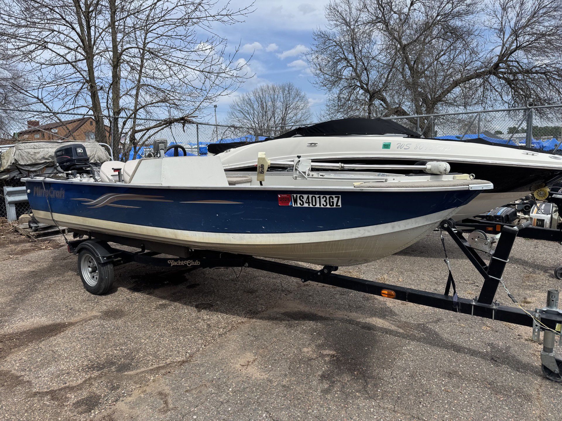 Blue-and-white boat on a trailer in a parking lot, with trees in the background.