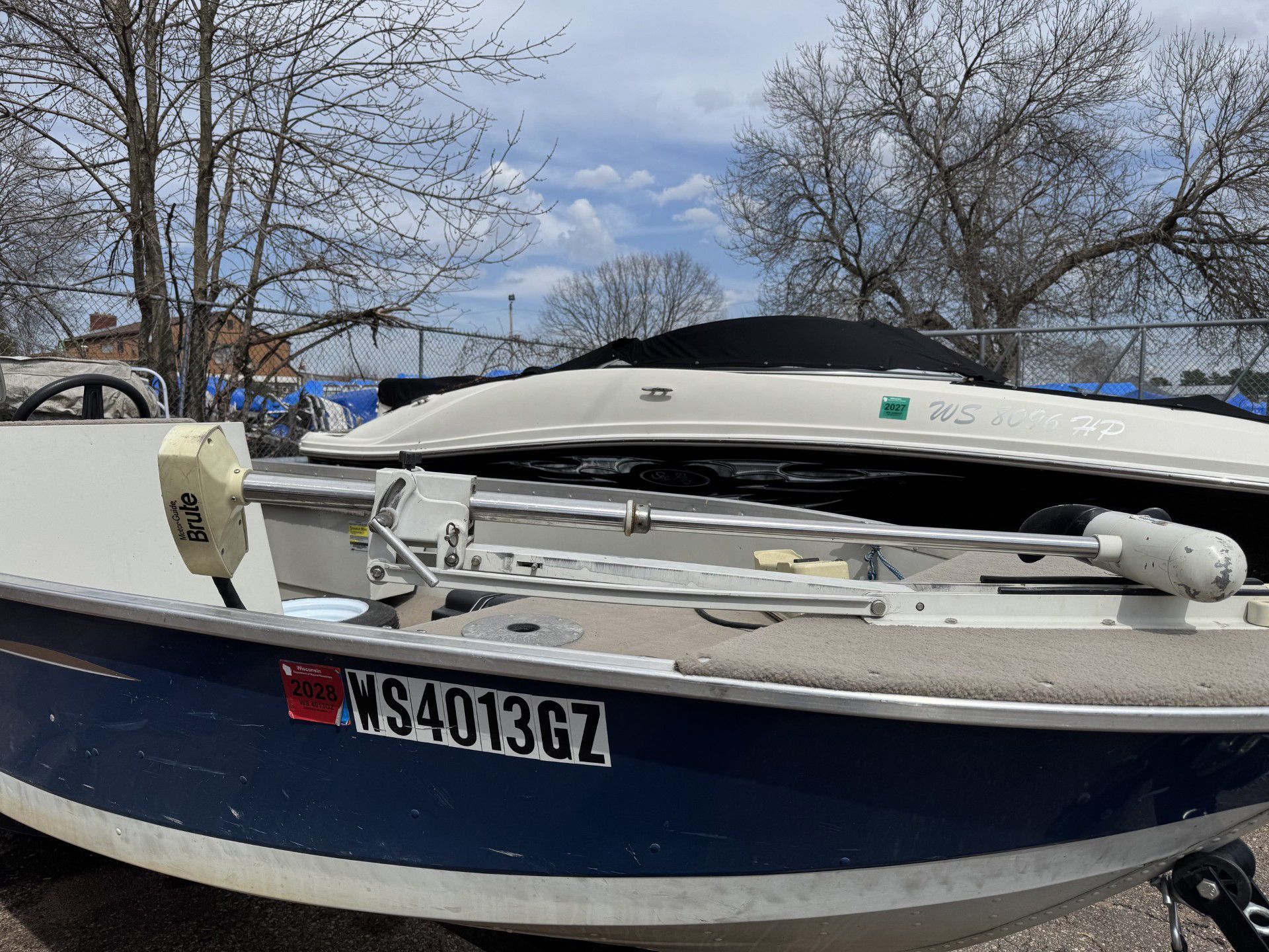 Blue boat with trailer and white kayak on top, parked outdoors under bare trees.
