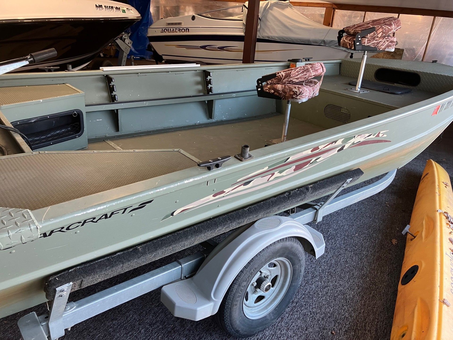 A small green aluminum boat on a trailer, inside a garage. Includes seats and fishing rod holders.