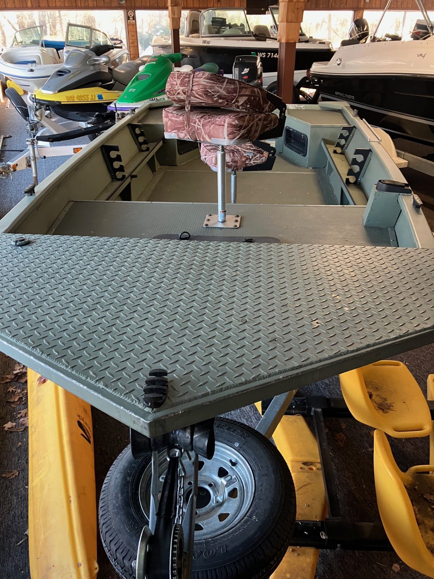 A green, flat bottomed boat on a trailer, with a camouflage seat, inside a building with other boats.