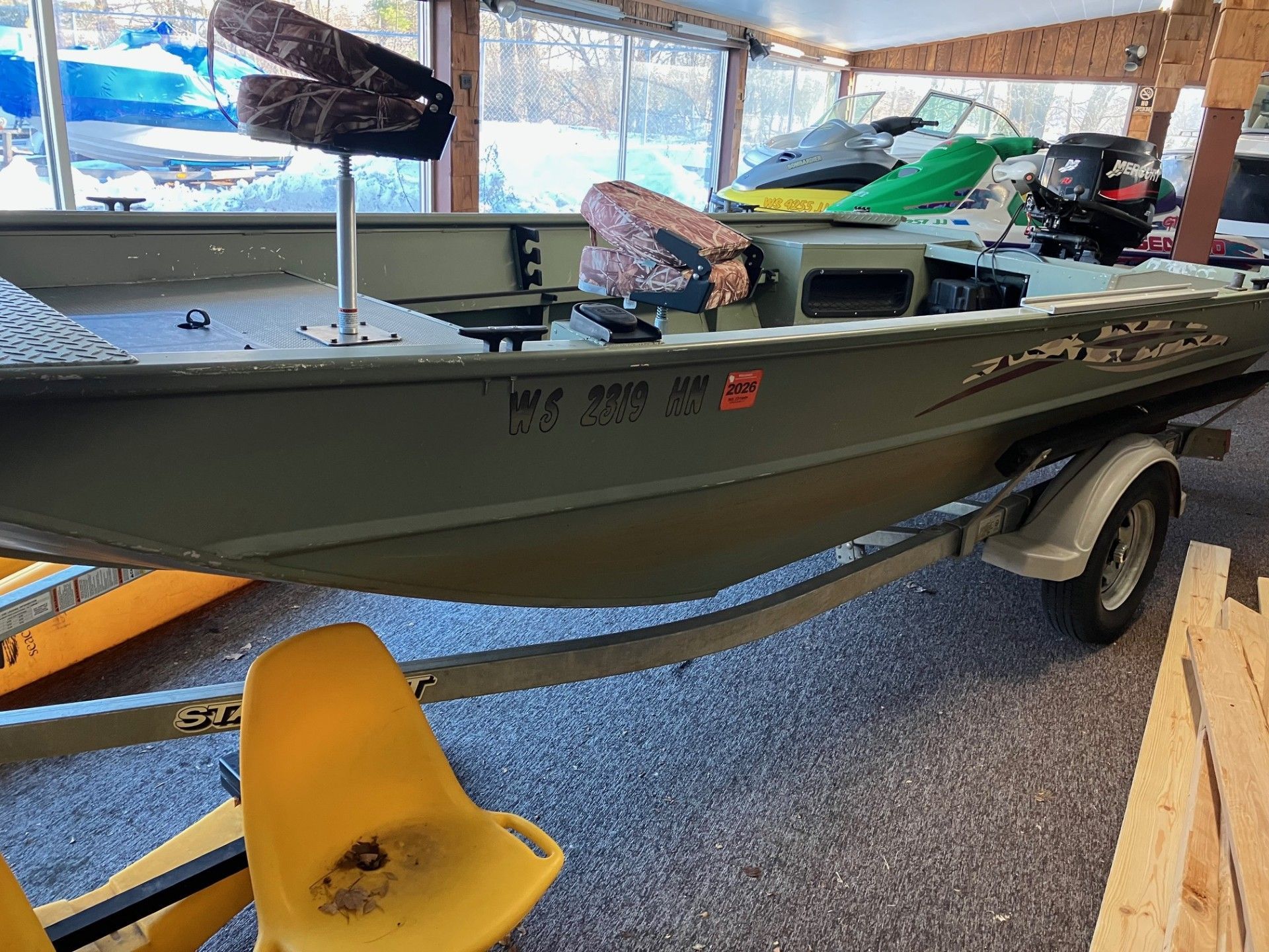 Green aluminum boat on a trailer, under a covered structure. Camo seats, WV registration, snow visible.