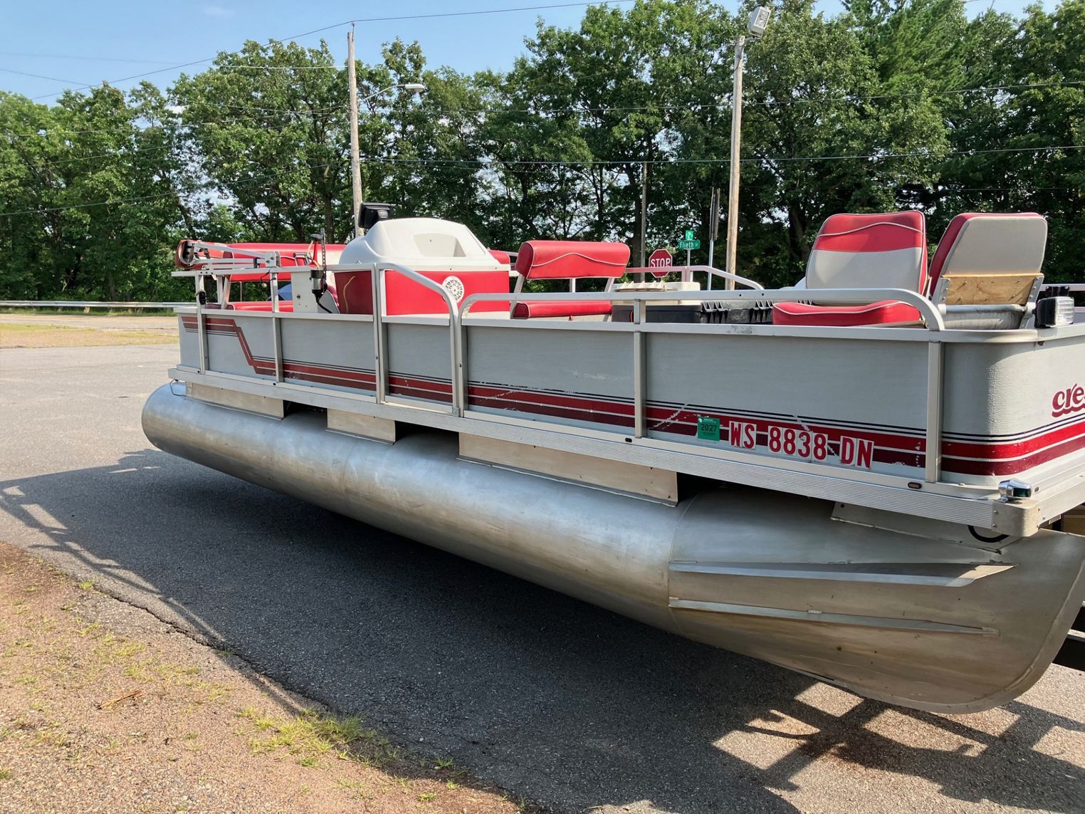 Pontoon boat on a trailer, light gray with red accents, parked on gravel. Trees in the background.