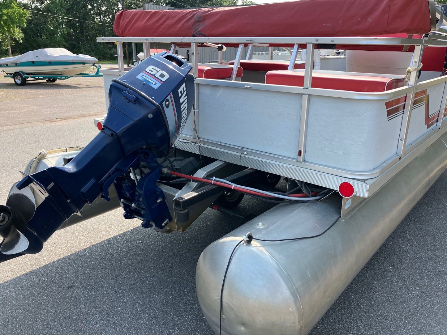 A pontoon boat with a blue motor sits on a paved area. It features red seating and trim, with another boat in the background.
