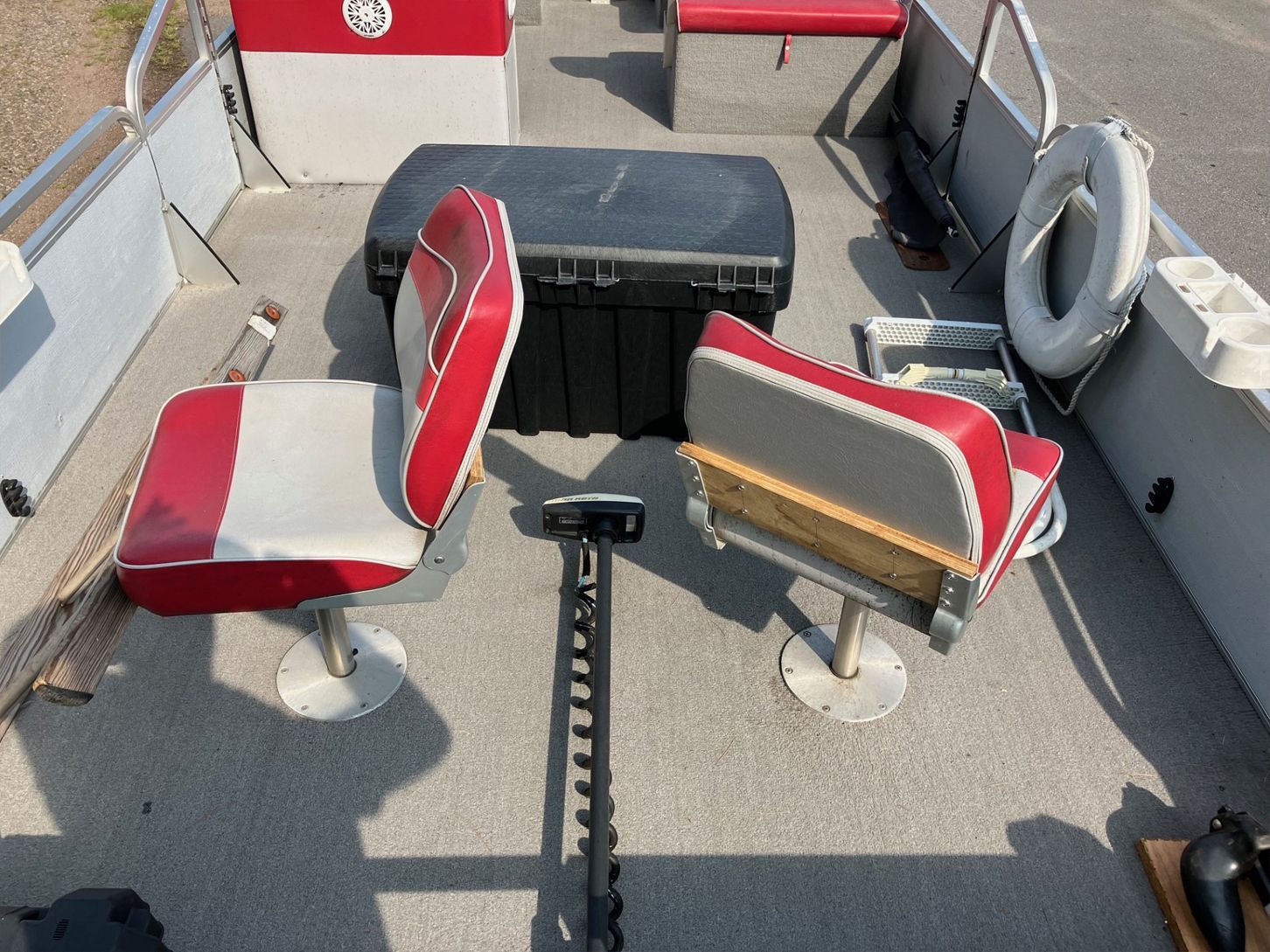 Inside a boat, two red and white seats face a black storage container and steering mechanism; other seating and a life preserver are visible.