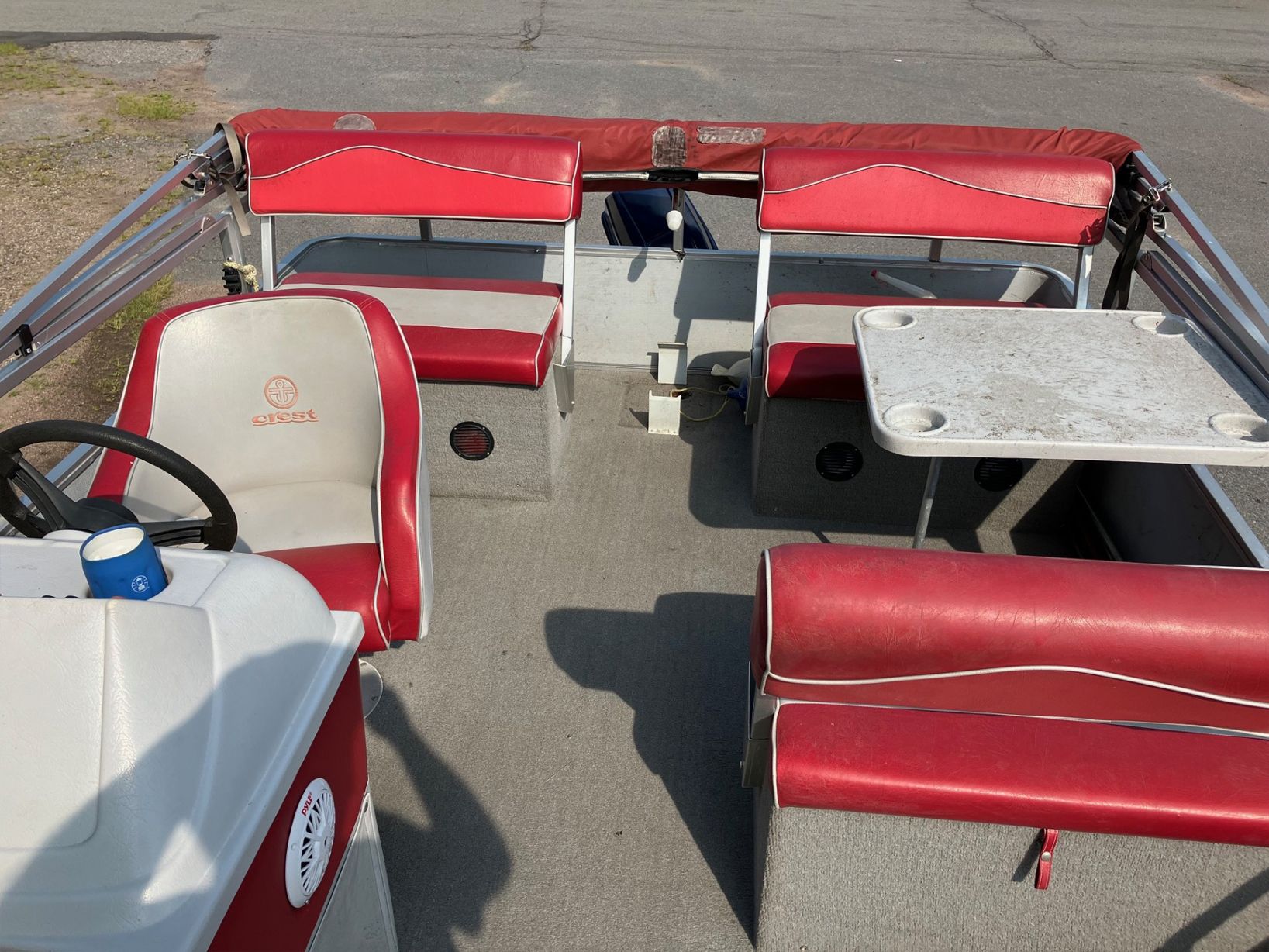 Interior of a pontoon boat with red and white seating, a table, and a steering wheel. The boat is on a paved surface.