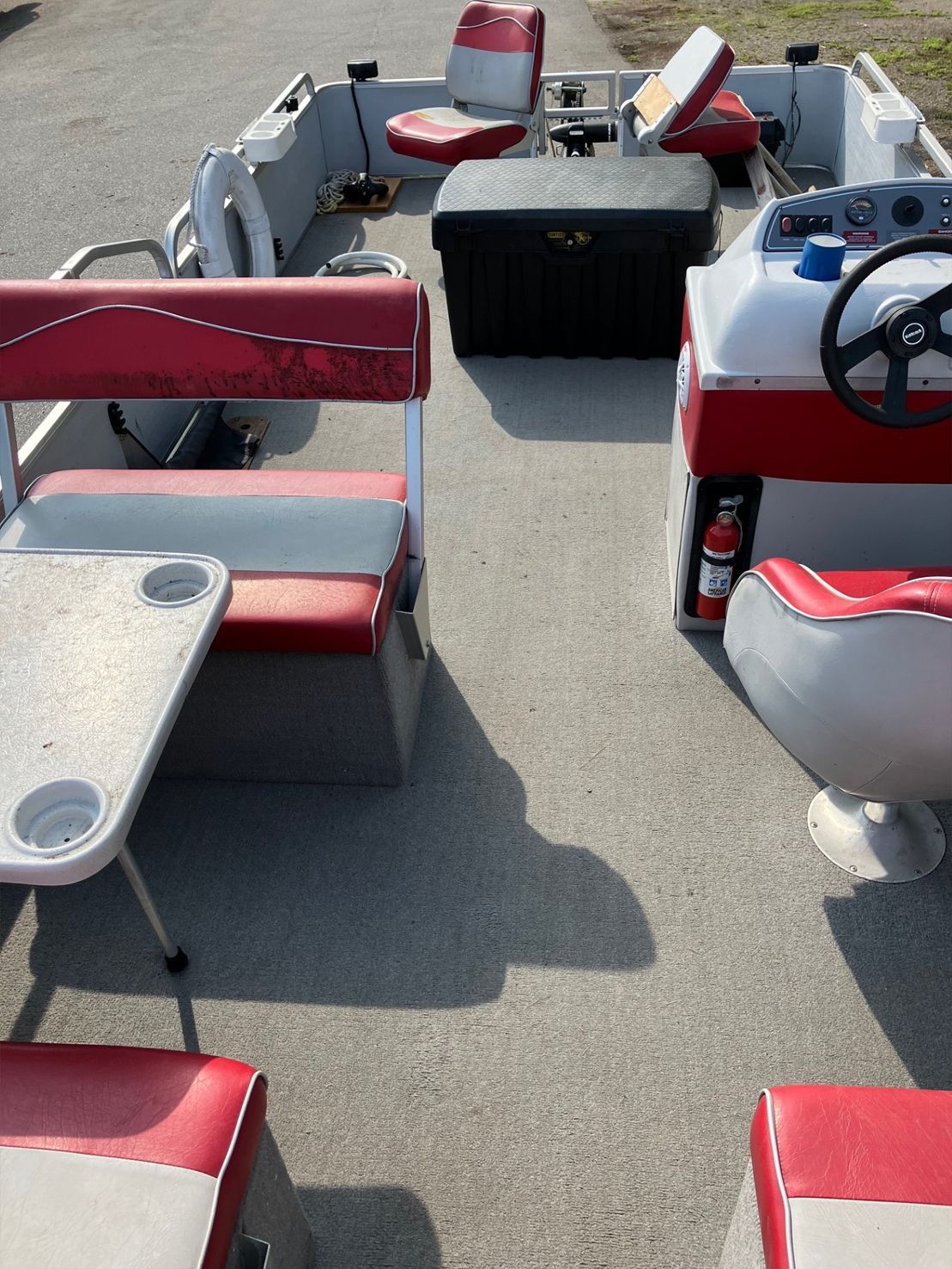 A boat's interior with red and white seating, a table, and a black storage container. The boat is in an outdoor setting.