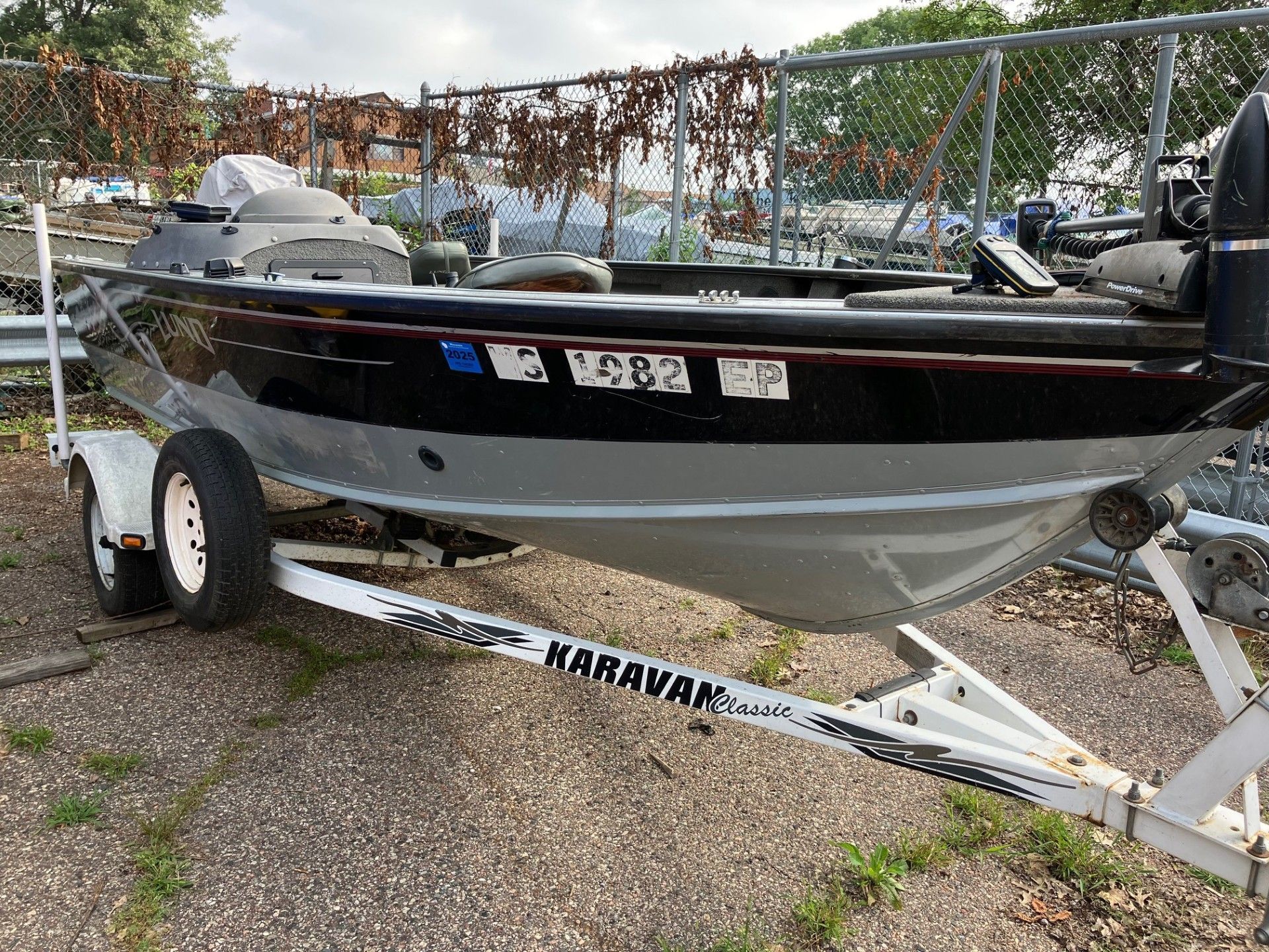 Boat on a trailer; aluminum hull with black and gray paint, parked in a grassy area near a fence.