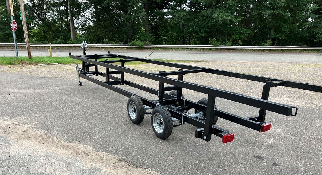 Black trailer with two sets of wheels, designed for transporting long objects. 