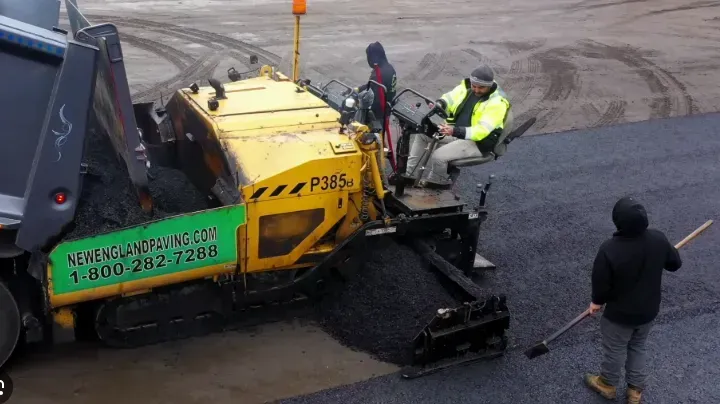 Asphalt paving in progress: workers operating a machine, receiving asphalt from a truck, laying it on a surface.