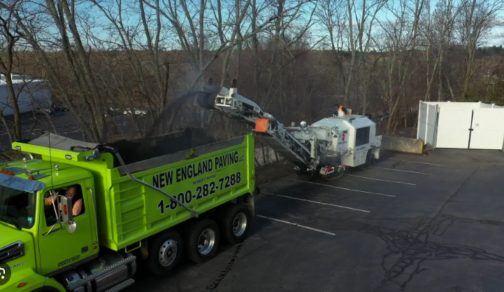 A road milling machine loading asphalt into a bright green dump truck in a parking lot.