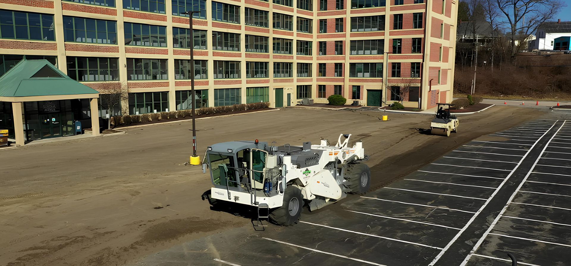Construction vehicles paving a parking lot in front of a brick building with many windows.