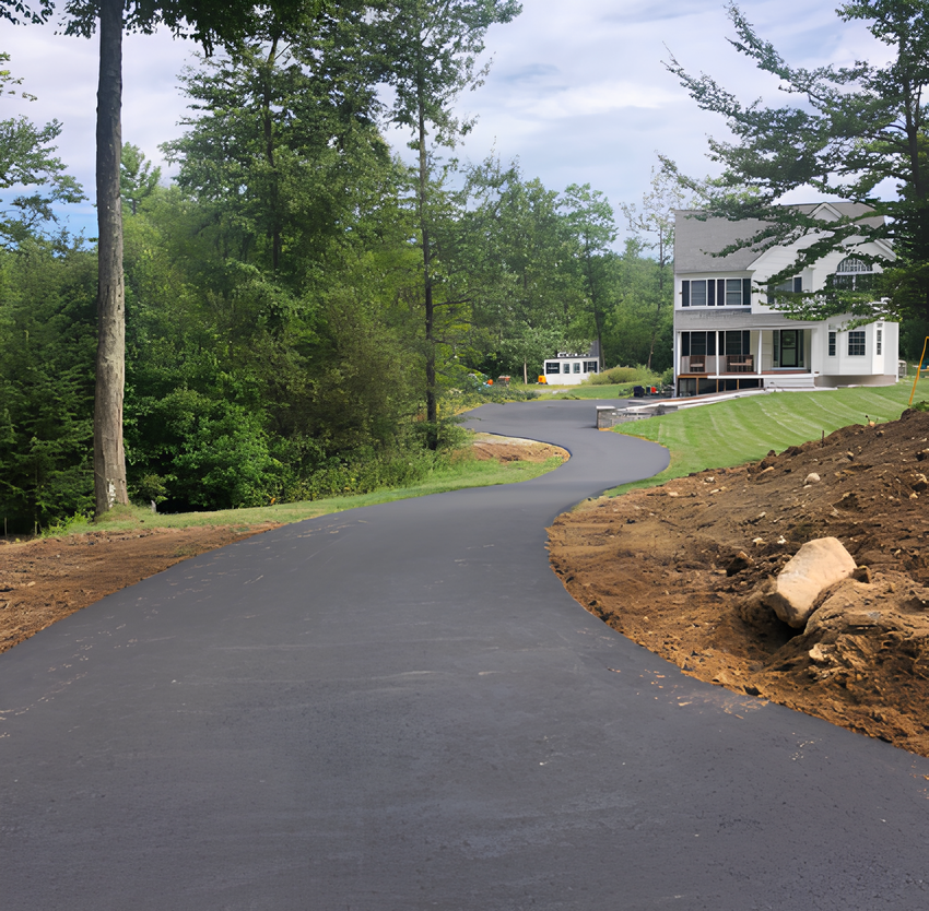 Paved driveway curves toward a two-story house, bordered by trees on one side and bare earth on the other.