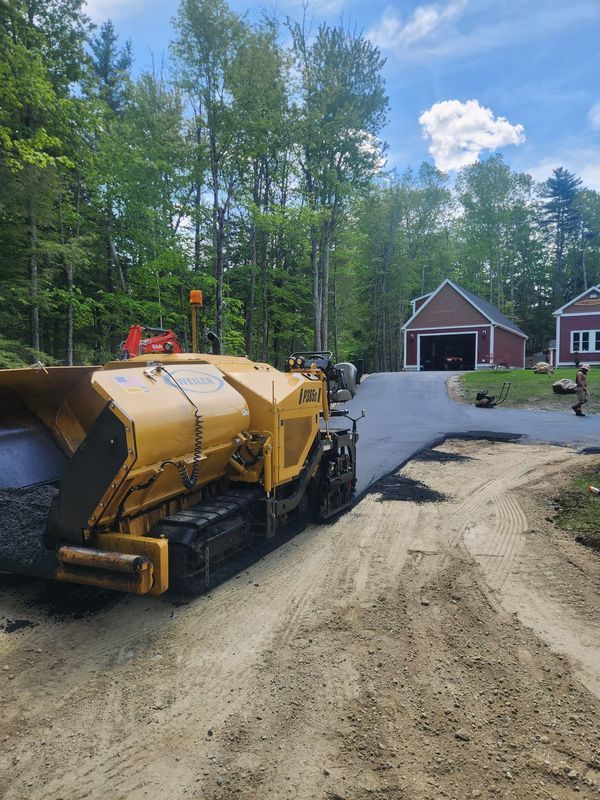 Yellow paving machine laying asphalt on a driveway, with a red garage in the background. Green trees and a blue sky.