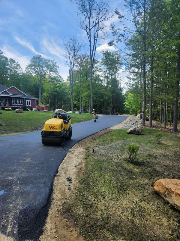 Asphalt driveway being rolled by a yellow compactor machine, trees and house in background.