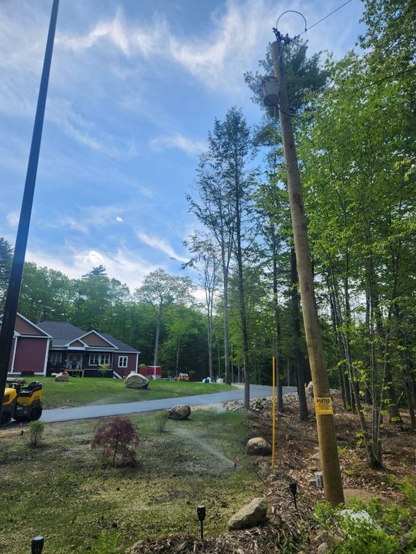 Power pole with transformer, house, trees, and sky. Sprinkler system watering grass.