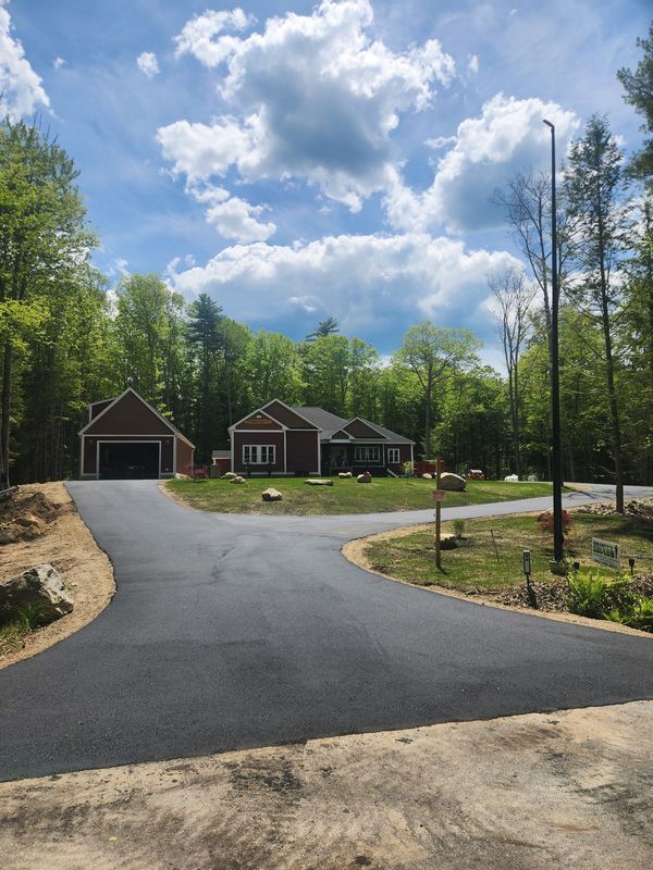 Driveway leading to a brown house and garage surrounded by trees and a blue sky with clouds.