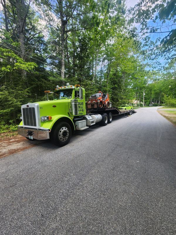 Green semi-truck hauling a small excavator on a trailer, parked on a road lined with trees.