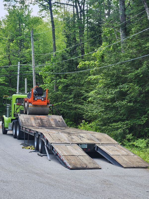 Green semi-truck hauling a roller compactor on a flatbed trailer along a tree-lined road.
