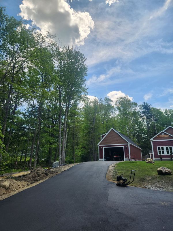 Paved driveway leading to a red garage and house, under a cloudy blue sky, with trees lining the sides.