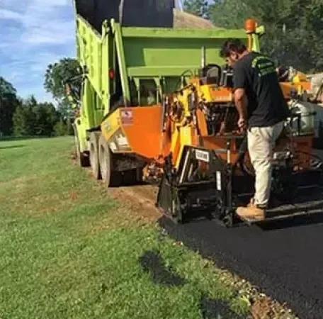 A worker operating asphalt paving equipment, next to a dump truck, laying asphalt on a road edge.