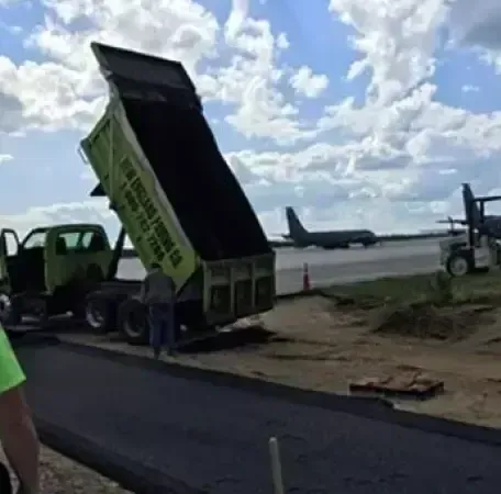 Truck dumping asphalt onto a road. Construction site with a plane in the background under a cloudy sky.