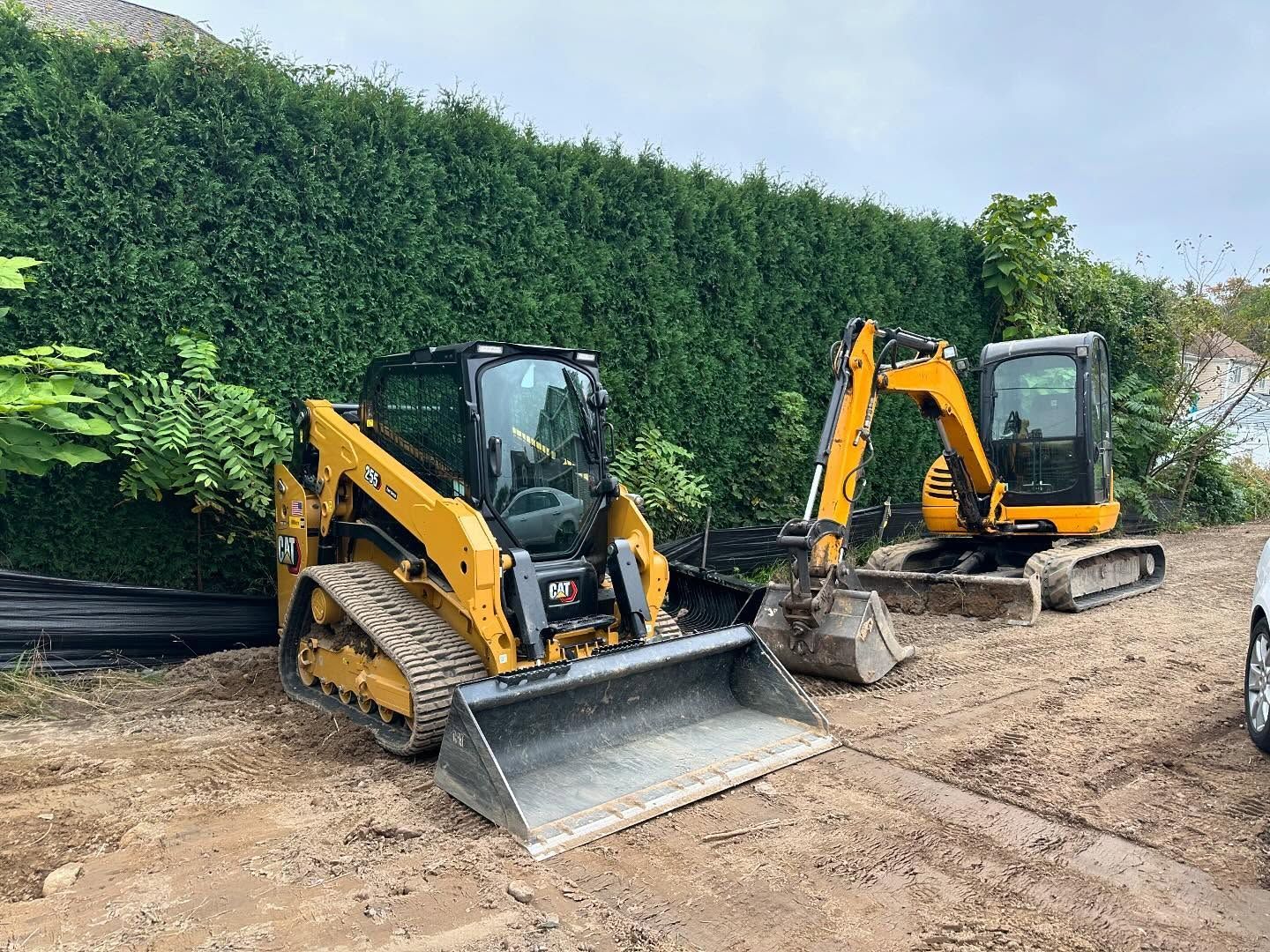 Two yellow skid steer loaders with buckets parked on a dirt lot in front of a tall green hedge