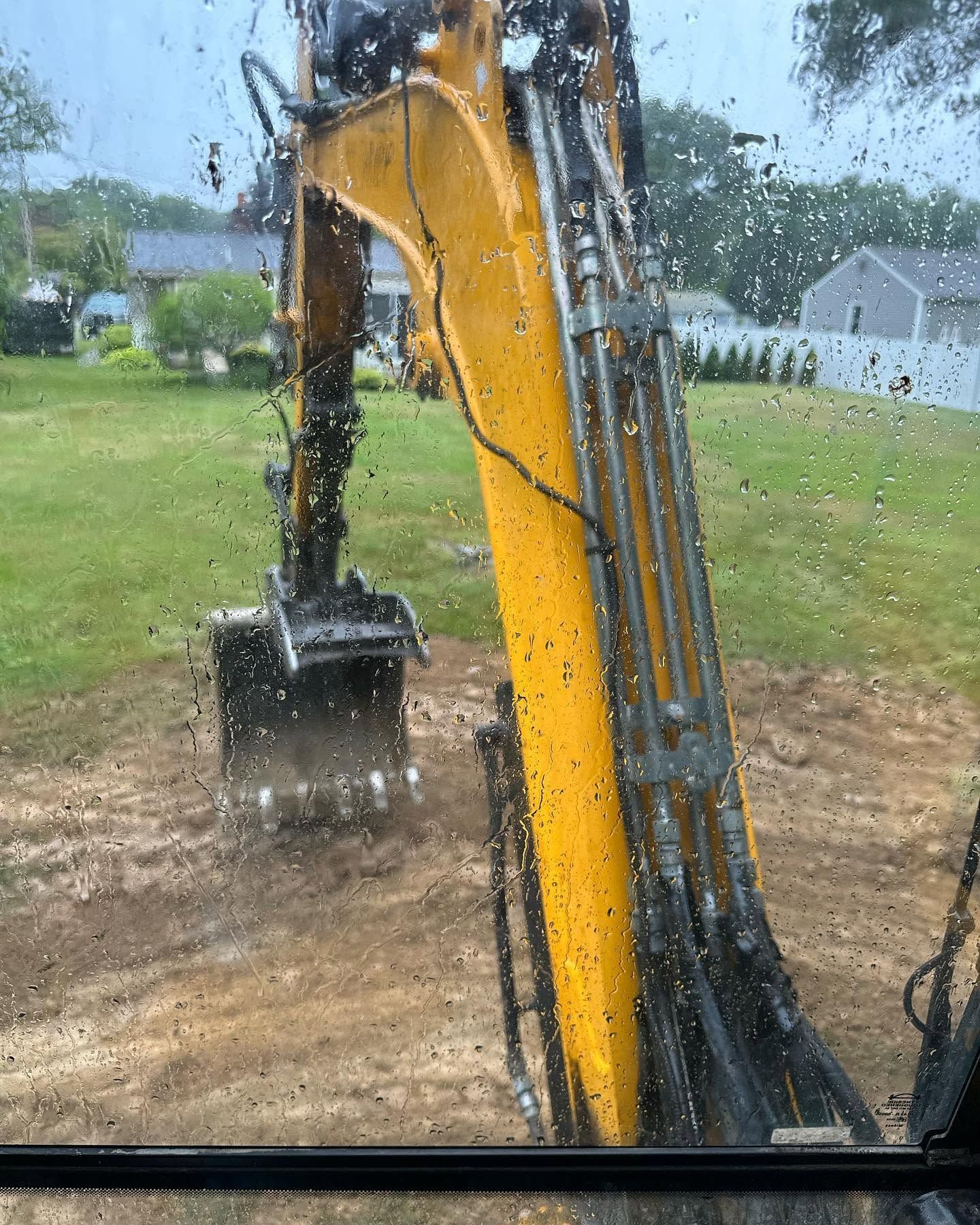 Yellow excavator arm and bucket digging dirt in a rainy yard, viewed through a wet window.