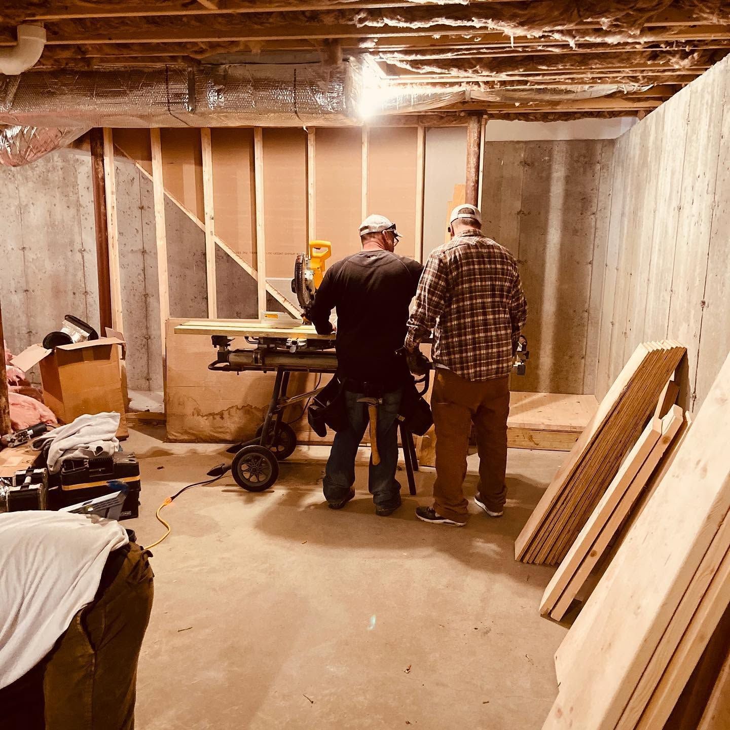 Two people working in an unfinished basement near a table saw and stacked lumber under exposed beams