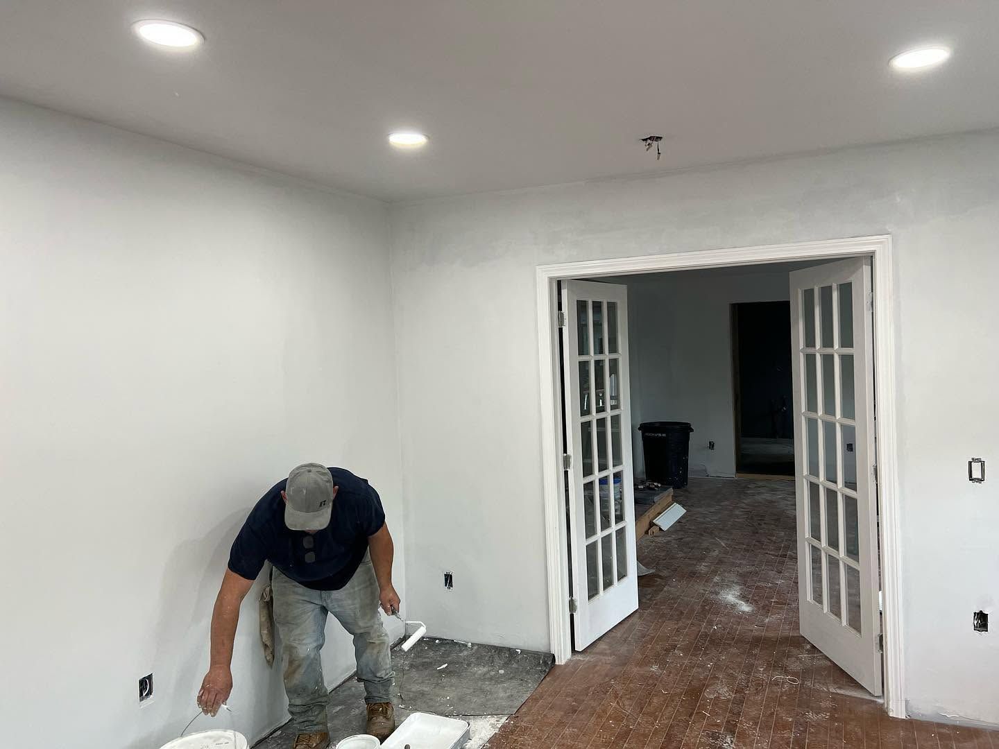 Person installing flooring in a white room with recessed lights and open double doors.