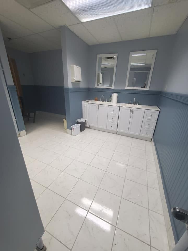 Small tiled restroom with blue-gray walls, white sink cabinet, mirror windows, and a trash can.