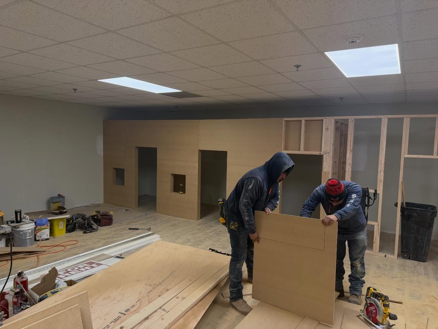 Two workers framing interior walls in a room under construction with lumber, tools, and drywall stacked nearby