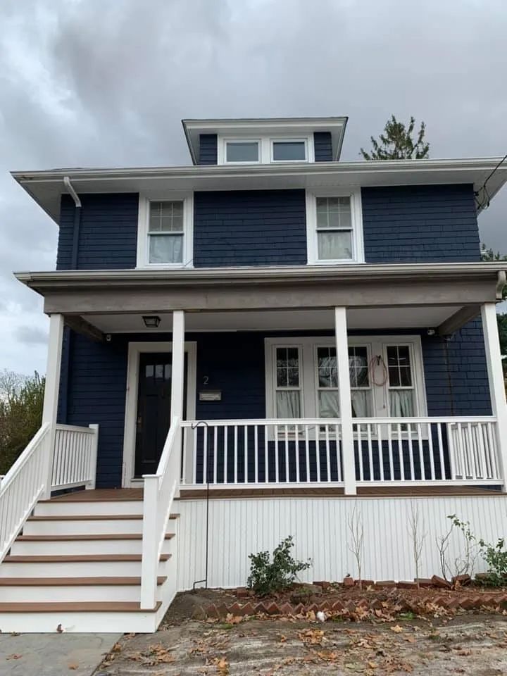 Blue two-story house with white trim, front porch, stairs, and dormer windows under an overcast sky