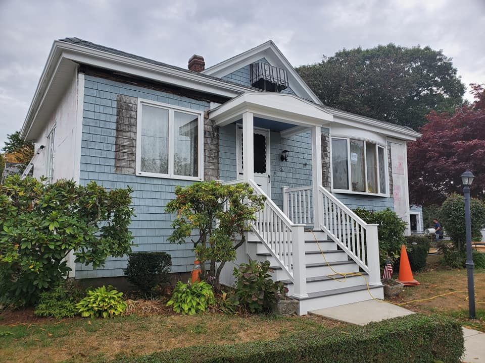 Blue house with white porch steps, front garden, and orange cones on a cloudy day