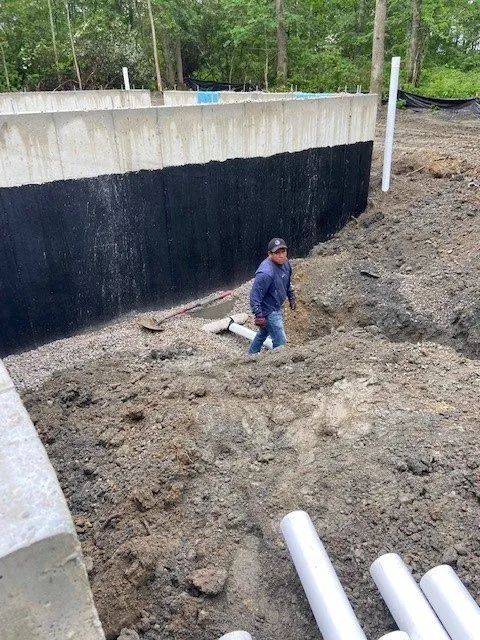 Worker standing in a dirt excavation beside a concrete wall and white pipes