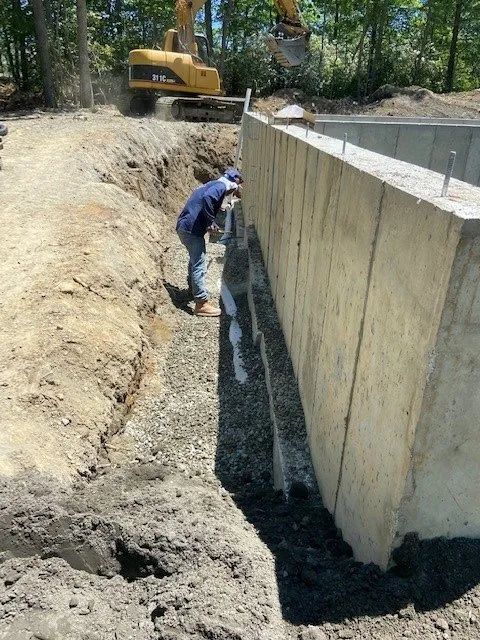 Worker inspecting a deep concrete trench beside a retaining wall, with an excavator in the background.