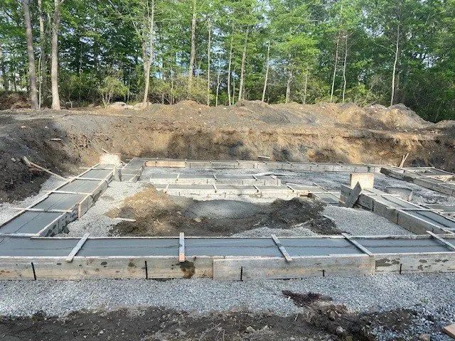 Concrete foundation forms for a house under construction, surrounded by gravel and trees.