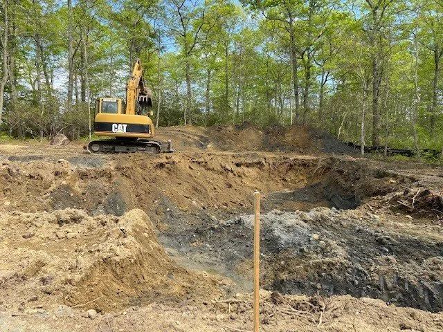 Excavator on a dirt site beside a wooded area with exposed earth and a marked stake in the foreground