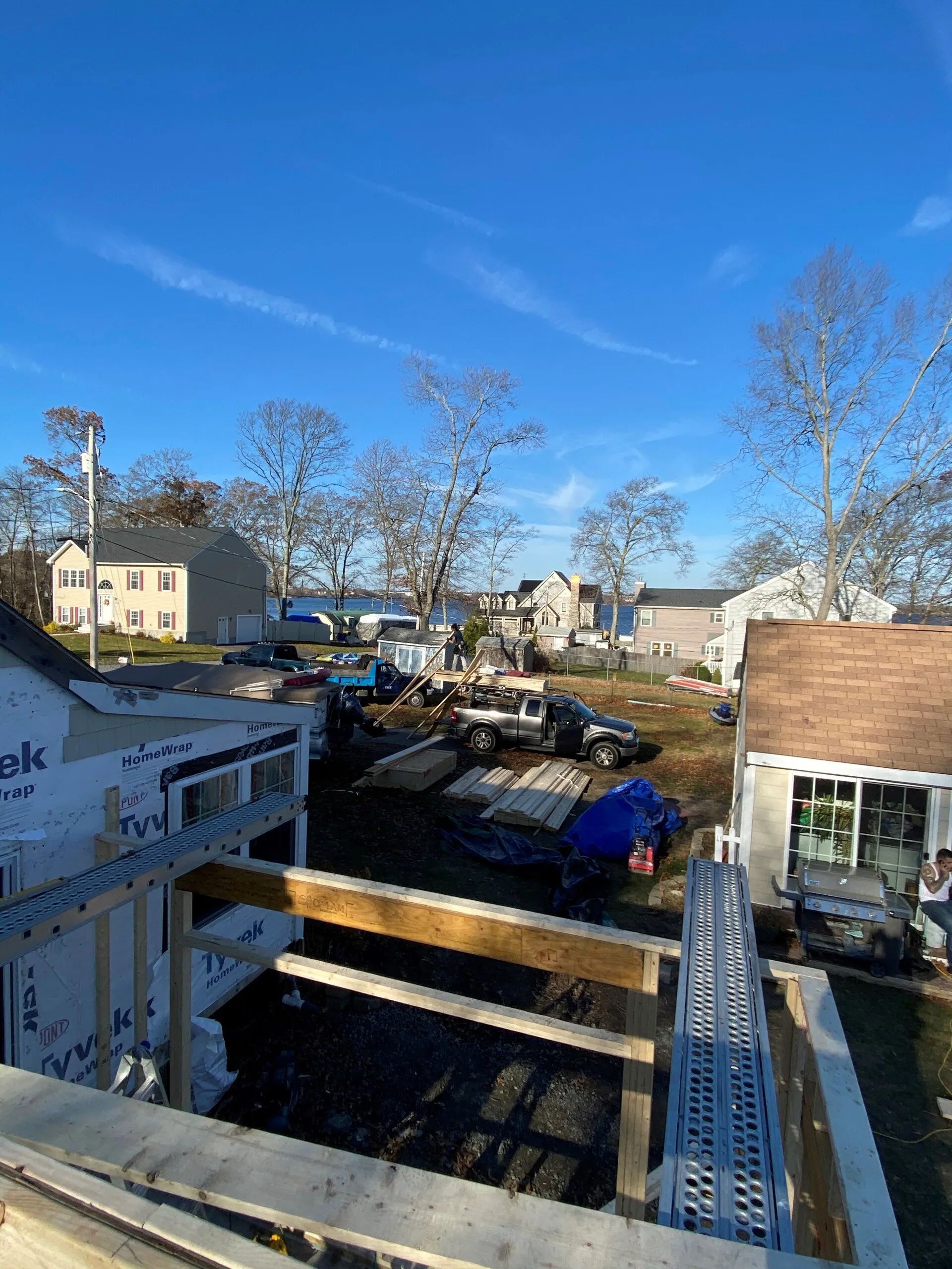 House under construction with exposed wooden beams, scaffolding, and neighboring homes under a clear blue sky
