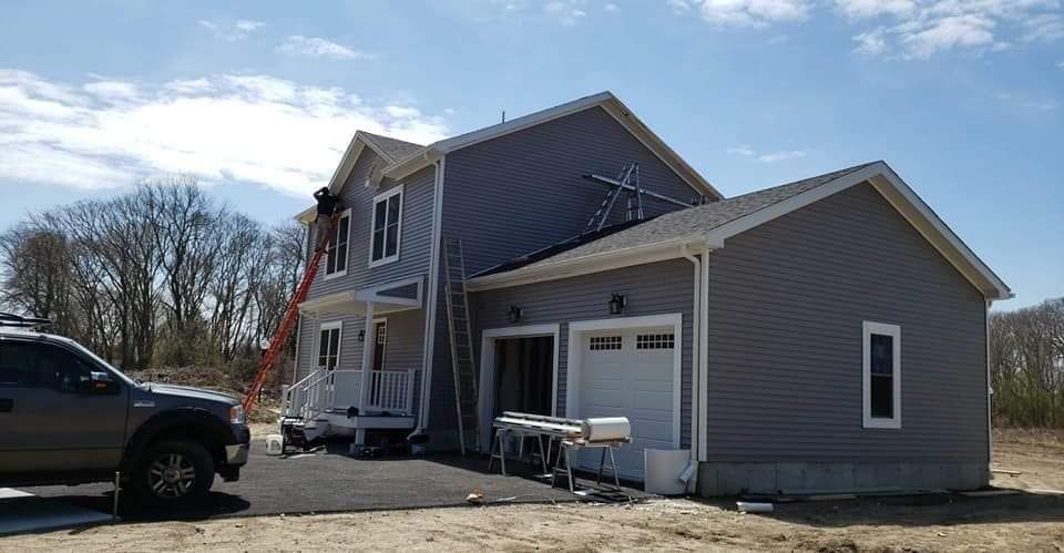 House under construction with workers on a ladder, gray siding, and a parked pickup truck outdoors
