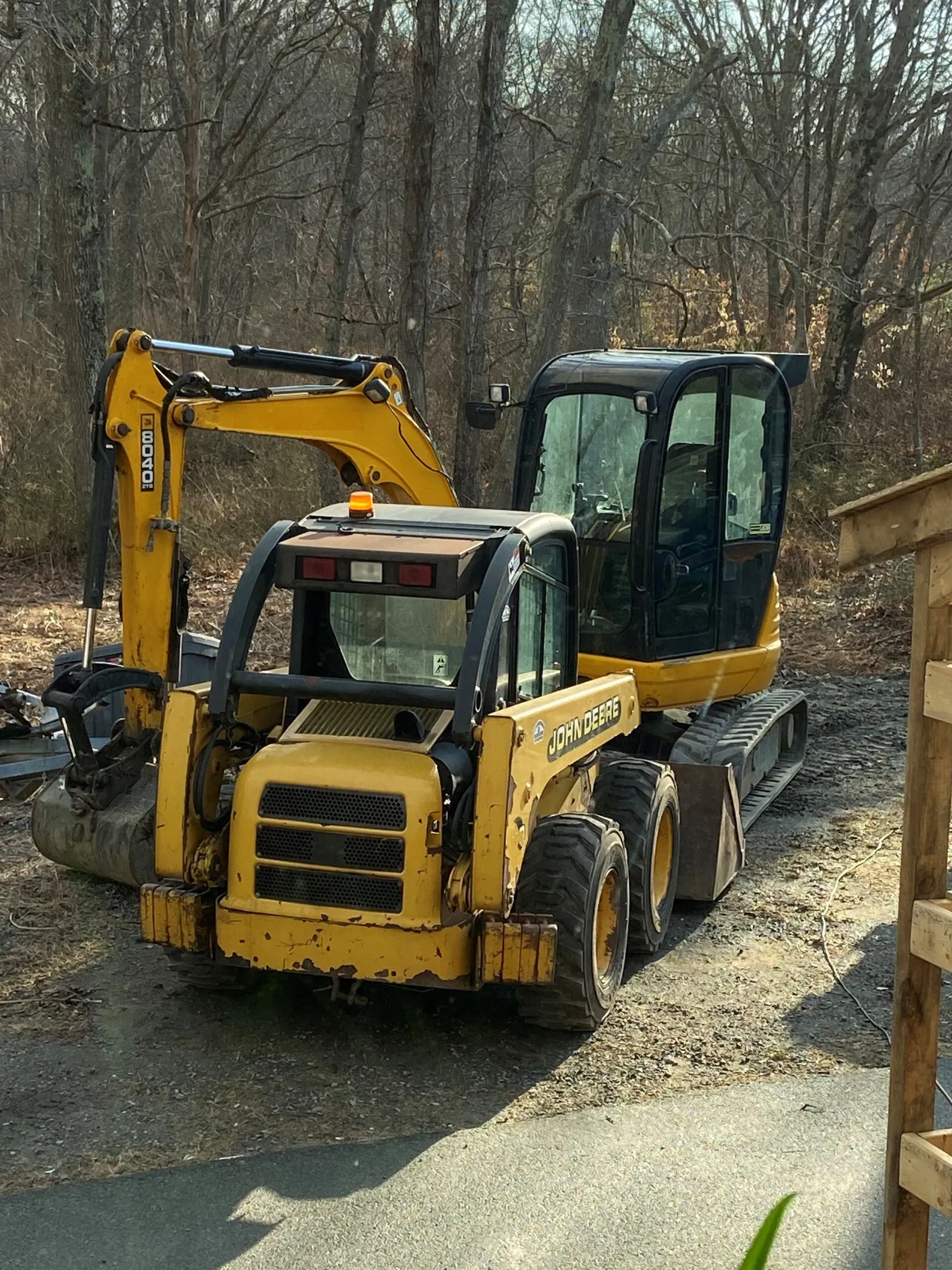 Yellow compact excavator on a muddy wooded worksite, with a larger excavator behind it.