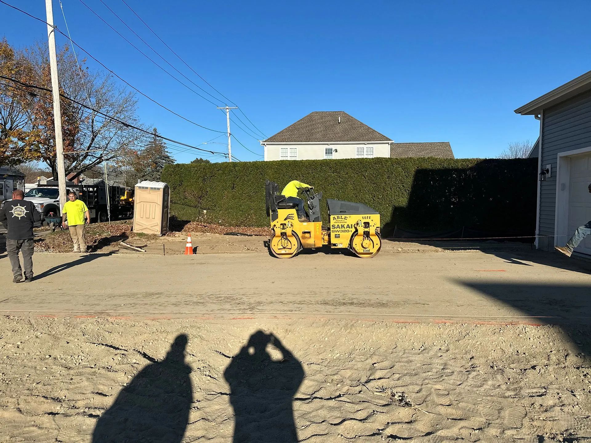 Construction site with yellow loader, workers, hedge, cones, and shadows on dirt ground