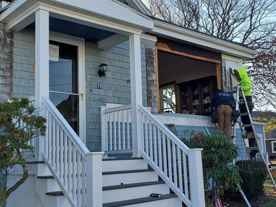 Worker repairs porch of a blue house, using a ladder beside the front steps.