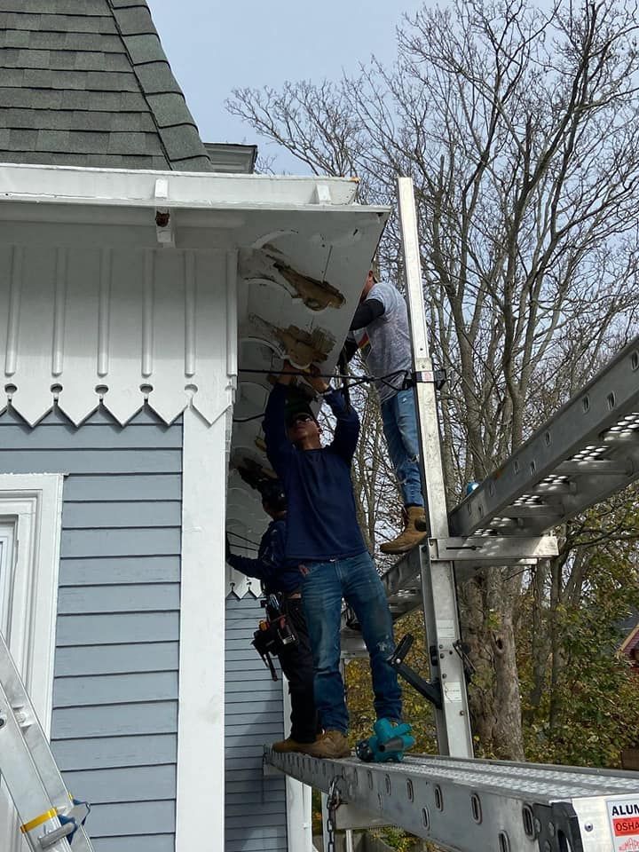 Two workers repairing a white house roof edge on scaffolding, with one holding damaged siding overhead.