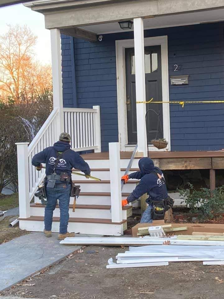 Workers installing a white accessibility ramp outside a blue house, with lumber and tools nearby