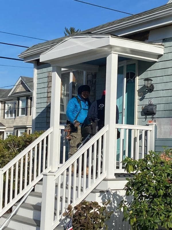 Person in a blue jacket standing on a front porch with white railings and steps.